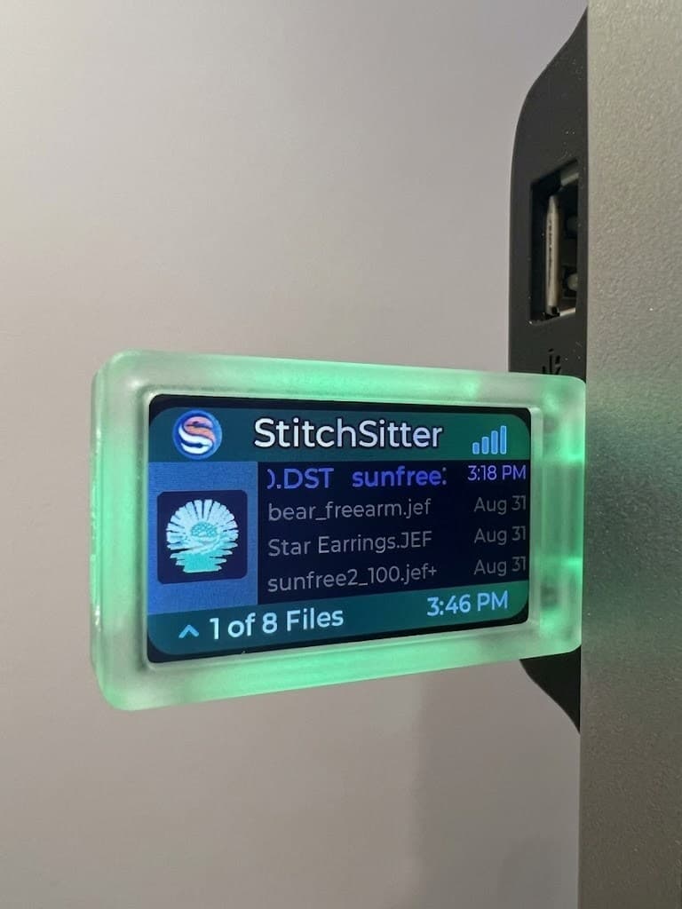 A StitchSitter dongle lit green, plugged into an embroidery machine's USB port, its screen listing the designs on the drive